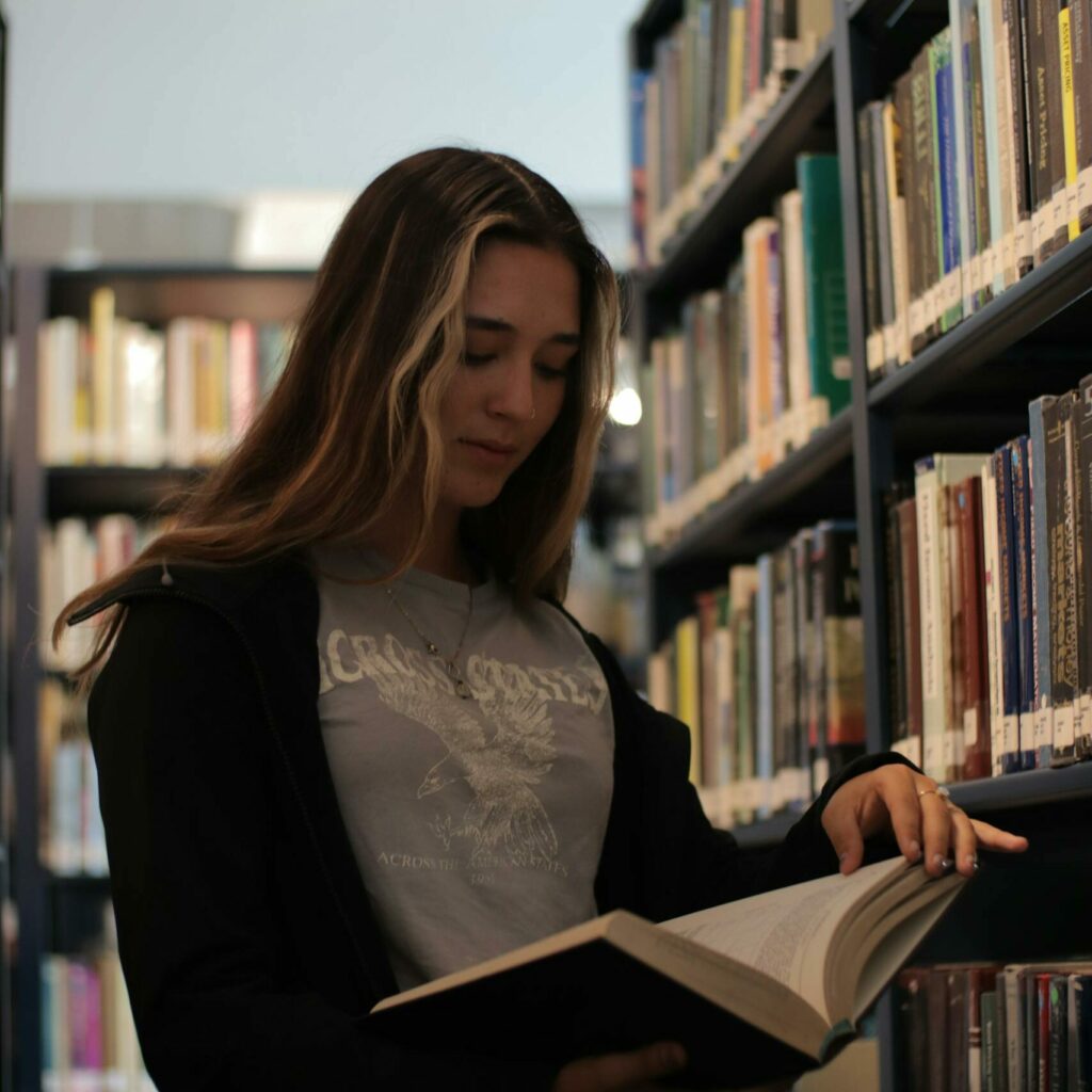 A young woman reading a book in a calm library setting, surrounded by shelves.